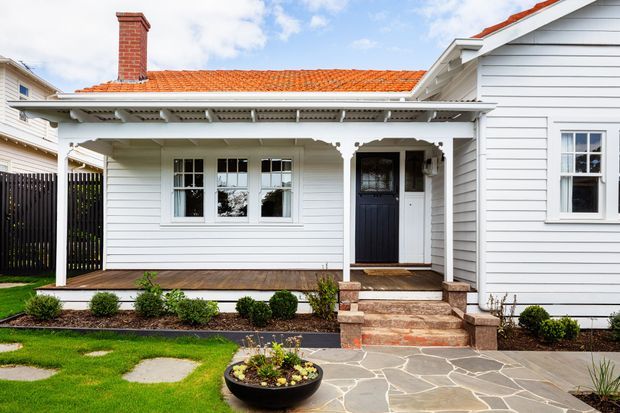 Front exterior of a traditional single-storey weatherboard house featuring a covered veranda with decorative timber fretwork, white-framed windows, a dark entry door, red terracotta tiled roof, brick chimney, brick steps, and irregular stone paving with landscaped garden beds.