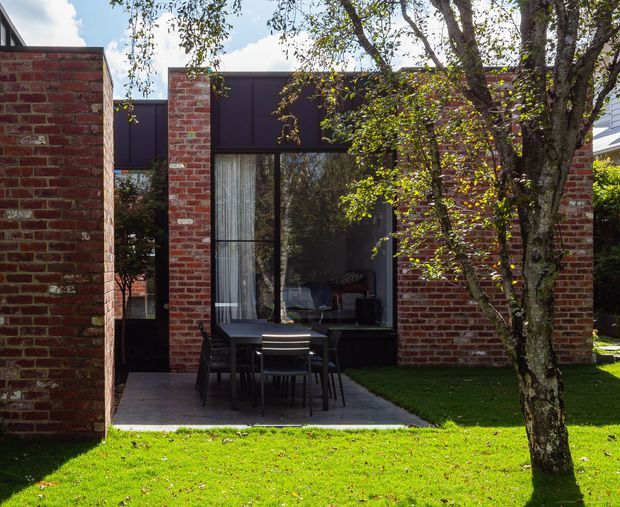 A single-storey residential extension or pavilion framed in exposed red brick, with dark metal cladding and large floor-to-ceiling glazing. A compact outdoor dining setting sits on a paved terrace in front of the window, surrounded by a manicured lawn and mature trees. The architecture has a strong indoor-outdoor connection and a restrained contemporary character.
