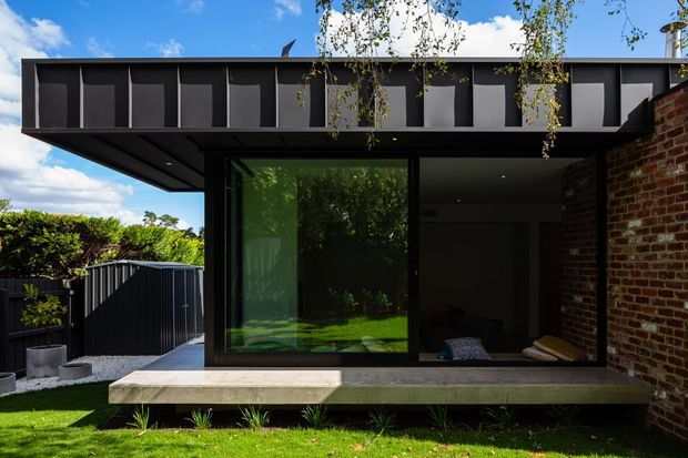 Exterior view of a contemporary flat-roof extension with a dark metal-clad upper facade, deep roof overhang, large black-framed glazed openings, and an exposed red-brick side wall. The building opens onto a raised concrete terrace overlooking a small landscaped lawn and fenced courtyard.