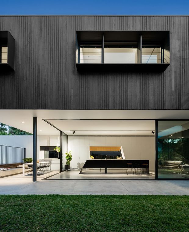 A contemporary two-storey house facade viewed from the garden, featuring dark vertically ribbed cladding on the upper level, deep-set projecting window boxes, and a ground-floor open-plan kitchen with extensive black-framed glazing. The kitchen has a large black island, pale tiled walls, timber cabinetry accents, and a covered outdoor terrace with dining furniture. A lawn and planted surroundings are visible in the foreground and through the glazing.