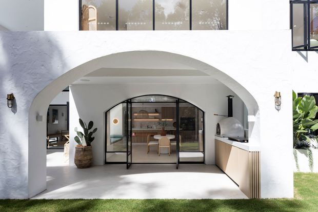 A bright white stucco courtyard-style exterior with a large rounded arch framing a glazed, black-framed kitchen or entertaining area. The space includes warm timber cabinetry, open shelving, a compact island or table, integrated appliances, a potted plant, and decorative wall sconces. A second-storey balcony or glazing is visible above, with landscaped lawn and tropical planting around the structure.