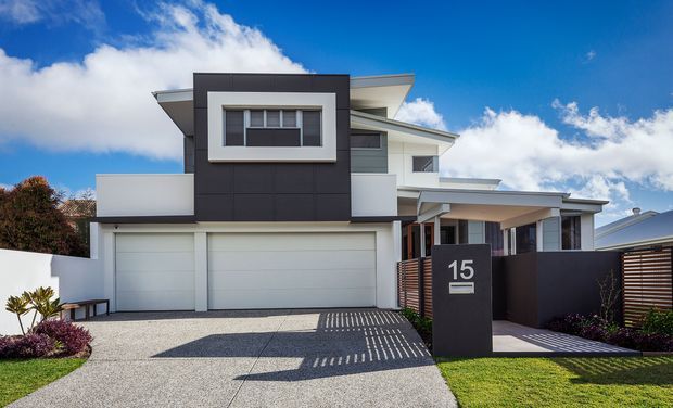 Street-facing view of a contemporary multi-level residence with a strong geometric facade, dark charcoal feature cladding, white rendered walls, broad eaves, upper-level glazing, an integrated double garage, and a screened entry courtyard. The property includes a textured concrete driveway, front landscaping, and a dark freestanding address pillar.