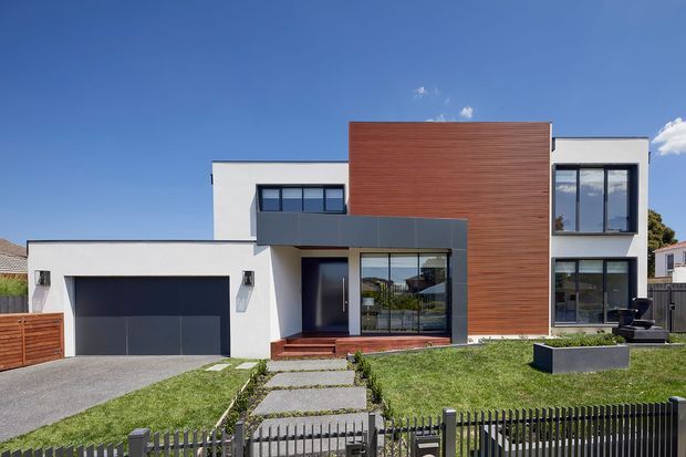 Front exterior view of a contemporary multi-level house with a crisp white rendered facade, prominent vertical timber-clad central volume, dark aluminium-framed glazing, an integrated double garage, paved entry path, lawn, and a black metal front fence under a clear blue sky.