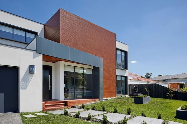 Street-facing view of a contemporary multi-level residence with a crisp geometric form, white rendered walls, dark-framed glazing, a prominent red brick feature volume, and a projecting grey metal balcony. The landscaped front yard includes a paved path, lawn, low planting, raised planters, and a dark fence. A garage and neighbouring homes are also visible.