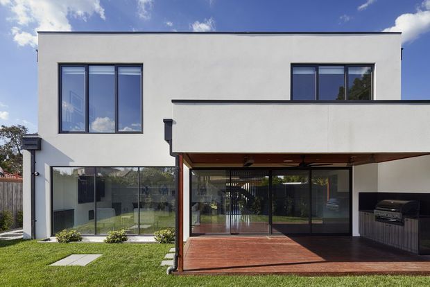Contemporary two-storey white-rendered house viewed from the rear, with dark-framed windows and extensive ground-floor glazing. A projecting upper-level terrace or balcony forms a covered outdoor entertaining area with a timber deck and built-in outdoor kitchen. The landscaped lawn and clear glazing create a clean, modern residential facade.