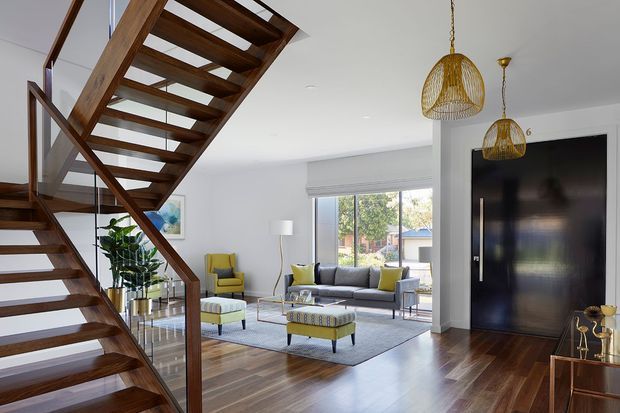 Bright contemporary open-plan living room viewed from beside a sculptural open-riser timber staircase. The room features polished dark timber flooring, white walls and ceiling, a grey sofa with yellow cushions, matching yellow occasional chairs and ottomans, large windows with roller blinds, woven pendant lights, and a dramatic dark built-in wall or door element.