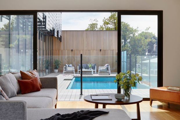 A contemporary living room viewed through broad dark-framed glazing toward a private outdoor pool courtyard. The room includes a large light-grey sofa, round timber coffee table, low timber console, and warm timber flooring. Outside, a compact blue swimming pool is enclosed by glass balustrades and a vertical timber privacy screen, with outdoor lounge chairs beyond.