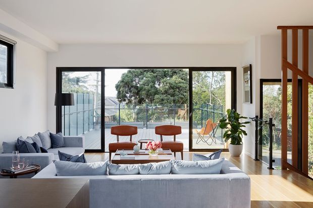 Bright contemporary living room with a large pale sectional sofa facing a glass coffee table and low console, blue-grey accent cushions, and a pair of terracotta lounge chairs. Full-height black-framed glazing opens to a balcony or terrace with landscaped views. Light timber flooring, white walls, a black floor lamp, indoor greenery, and a timber stair balustrade define the open-plan space.