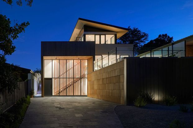 Twilight exterior view of a contemporary multi-level residence, showing a paved driveway leading to a tall glazed garage or entry volume with vertical timber screening. The home features dark metal cladding, warm timber elements, extensive glazing, a projecting upper roof, and landscaped boundaries.