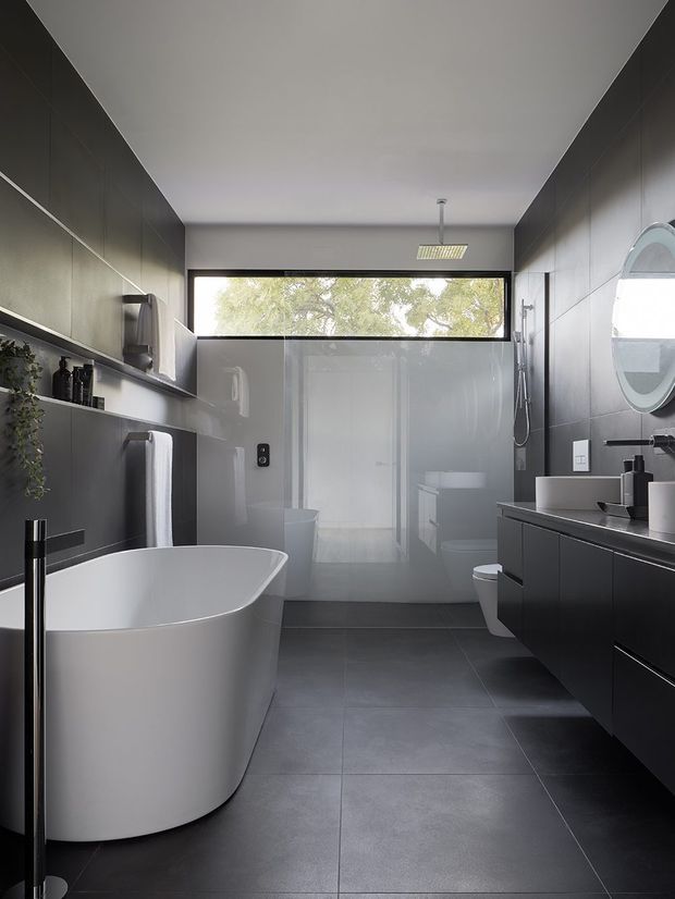 A contemporary bathroom with a freestanding white bathtub, wall-mounted dark vanity, integrated basin, round mirror, walk-in shower, and toilet. Large dark grey floor and wall tiles create a streamlined monochromatic scheme, while a high horizontal window provides natural light and views of greenery. Fixed shelving and towel storage line the left wall.