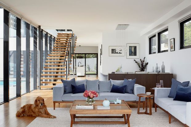 Bright contemporary open-plan living room with floor-to-ceiling glazing, a timber-and-metal open staircase, pale timber flooring, and a large neutral sectional seating arrangement. Navy cushions and a floral arrangement add colour, while a timber coffee table and dark wood sideboard provide contrast. The room opens toward a dining or kitchen area and has views to an outdoor pool area through the glazing.