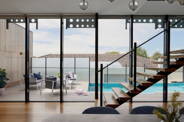 Contemporary open-plan interior with warm timber flooring and extensive black-framed floor-to-ceiling glazing overlooking a swimming pool and outdoor seating area. A floating timber staircase with dark metal stringers and balustrade occupies the right side, while a dining table and chairs sit in the foreground. The room is bright and visually connected to the outdoor pool area.