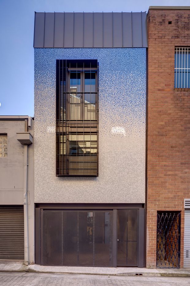 Street-facing view of a narrow contemporary urban building facade. The facade features a pale blue-and-white mosaic tile or patterned finish, a tall vertically proportioned glazed opening with dark metal security bars, dark metal-framed ground-floor doors, and a contrasting red-brown brick building immediately adjacent. The upper edge has a simple dark standing-seam metal parapet or cladding.