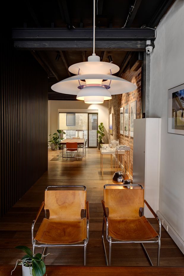 A narrow, stylish interior corridor or living area with two leather sling armchairs in the foreground beneath a large sculptural pendant light. The space features dark timber flooring, exposed dark timber beams and ceiling structure, a black vertical slatted wall, exposed red brickwork, white plaster walls, framed artwork, and a glimpse of a dining area with a table, chairs, plants, and glazing at the far end. The overall aesthetic is warm, refined, and industrial with mid-century furnishings.