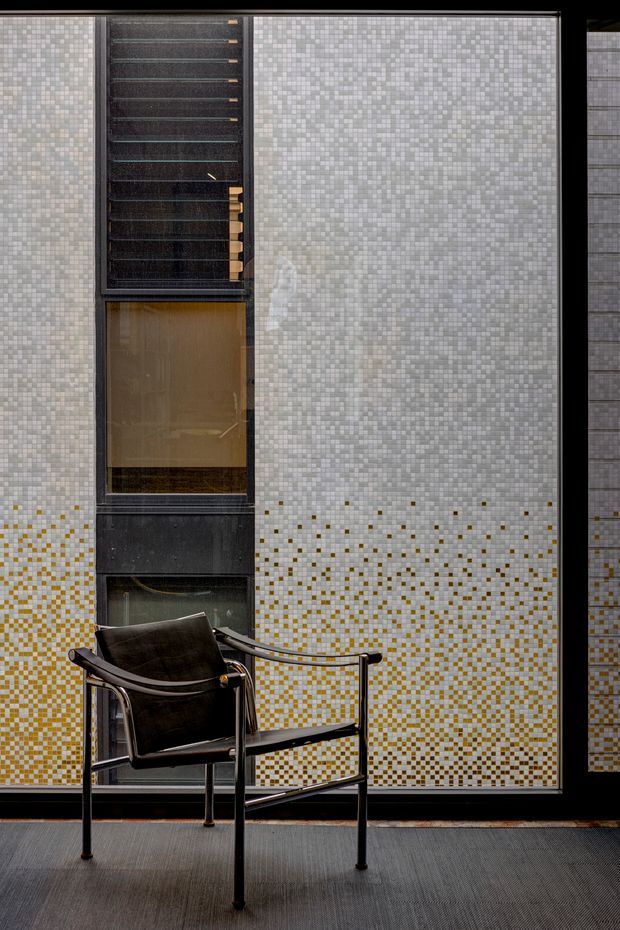 A close-up architectural interior view dominated by a large patterned mosaic-tile wall in pale grey and white, with a gradual field of mustard-gold accent tiles across the lower portion. A black-framed glazed opening and dark vertical architectural elements interrupt the wall. In the foreground is a minimalist black leather sling armchair with a tubular metal frame, set on a dark textured floor. The composition is refined and monochromatic with warm metallic accents.