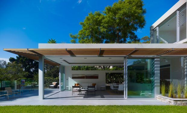 Exterior view of a contemporary modern home with a broad timber-lined flat-roofed outdoor entertaining pavilion connected to a glass-walled living area. The open-plan space contains outdoor-style lounge and dining furniture, while extensive glazing, slender metal frames, white walls, stone edging, and landscaped surroundings create a seamless indoor-outdoor connection. A swimming pool area is visible to the left.