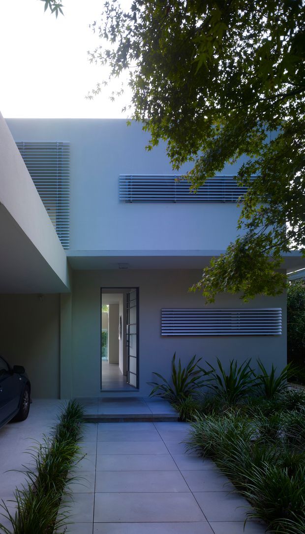 Exterior view of a modern, flat-roofed house entry. Pale rendered walls and horizontal metal louvre screens frame a recessed doorway, with a covered carport at left and a tiled concrete path leading through low ornamental planting. Mature tree foliage partially shades the facade.