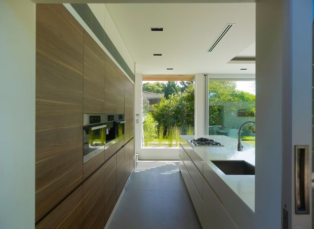A bright, contemporary galley kitchen with full-height dark timber-look cabinetry along one wall and a large pale island opposite. The island includes an integrated sink with a tall curved tap and a gas cooktop, while built-in ovens are set into the cabinetry. Floor-to-ceiling glazing at the end of the kitchen opens views to a lush garden, bringing strong natural light into the space. The ceiling has recessed lighting and linear air vents, and the floor is a large-format grey tile.