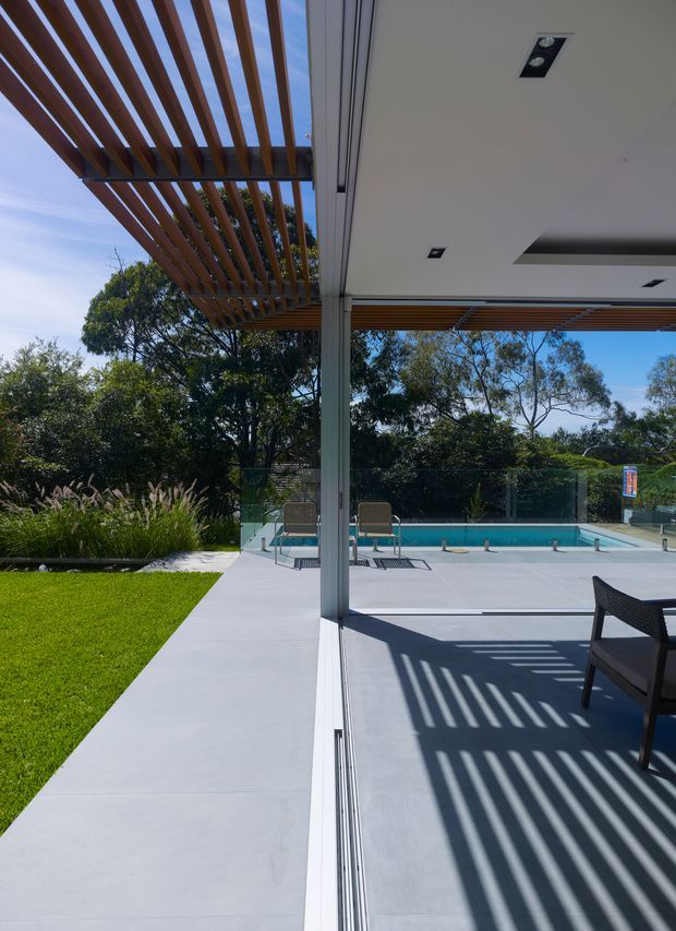 A bright contemporary indoor-outdoor entertaining area beside a swimming pool. The view looks across a pale concrete-paved terrace with a minimalist white ceiling, recessed lights, slim structural posts, and timber slatted pergola screens casting strong linear shadows. Glazed or openable doors connect the covered patio to the garden and pool deck, with mature trees and ornamental planting beyond.