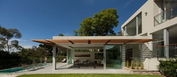 Exterior view of a contemporary multi-level residence with a broad timber-slat pergola extending over an outdoor entertaining area. The open pavilion contains a dining table and chairs, with large glazed walls connecting it to the house. A swimming pool and lounge area are visible to the left, while the main building has pale rendered walls, extensive glass, balconies, and horizontal privacy screens. Mature landscaping surrounds the property under a clear blue sky.