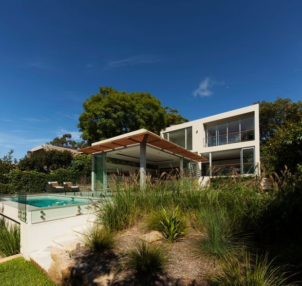 Exterior view of a contemporary multi-level residence with a crisp white rendered facade, extensive floor-to-ceiling glazing, projecting upper-level form, and a timber pergola shading an outdoor entertaining area. A turquoise swimming pool with glass safety fencing sits beside landscaped native-style planting and stone-edged paths. The image is captured in bright daylight from the garden.