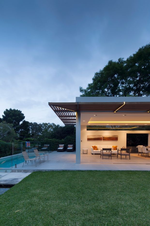 A dusk exterior view of a contemporary pavilion-like home opening onto a pool terrace and lawn. The structure has a flat roof with a timber slatted pergola, broad open glazing or openings, warm recessed ceiling lighting, and a minimalist indoor-outdoor living area furnished with sofas, chairs, and a dining table. A glass pool fence and swimming pool are visible to the left, with mature trees forming the background.