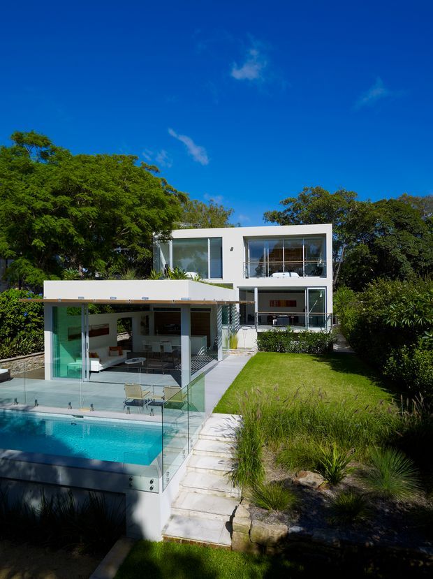 A bright contemporary two-storey white house set within a lush landscaped garden. The rear facade features extensive glazing, a covered outdoor living area, a balcony, and a turquoise swimming pool enclosed by frameless glass balustrades. Pale stone steps connect the lawn and pool terrace, with dense mature trees surrounding the property under a clear blue sky.