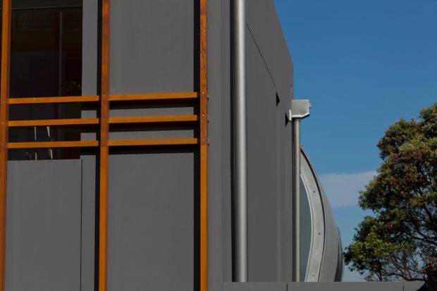 Close-up exterior view of a contemporary building corner, featuring dark grey wall cladding, warm timber-framed glazing and horizontal timber rails, a vertical metal downpipe, and a curved roof or facade element. A clear blue sky and tree are visible in the background.