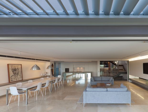 Open-plan contemporary living and dining area beneath a dramatic glazed roof structure. The room features a long dining table with white chairs, pale upholstered sofas, pendant and linear lighting, large-format light stone flooring, a central stone-clad or tiled feature wall, and a stairway with metal balustrade at the rear. The palette is light and neutral with strong exposed structural roof beams visible above.