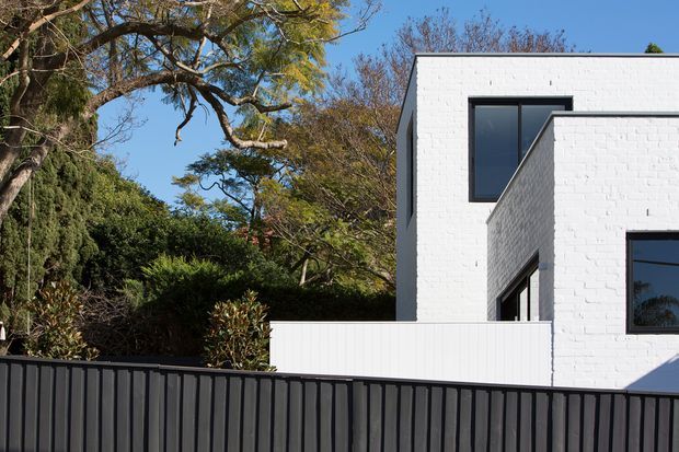Exterior view of a modern two-storey residence, featuring crisp white-painted brick walls, dark-framed rectangular windows, projecting geometric volumes, and a dark vertical-slat boundary fence. Established garden vegetation surrounds the building.