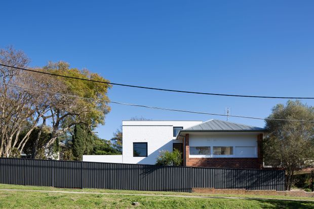 Street-facing view of a contemporary two-storey residence with a crisp white rendered upper volume, dark-framed square window, low-pitched grey metal roof, and a projecting brick-and-white glazed section. A tall dark metal privacy fence, mature trees, overhead utility wires, and a grass verge are visible in the foreground.