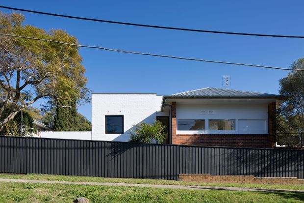 Street-facing view of a low-rise contemporary home behind a tall dark metal fence. The house combines a white rendered cubic section with a broad single-storey wing, pale metal hipped roof, large horizontal windows, and exposed red brickwork beneath the glazing. Mature suburban trees and overhead utility wires frame the property.