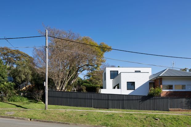 Street-facing view of a modern white two-storey residential extension or house behind a tall dark metal boundary fence. The building has a simple cubic form, flat roof, crisp white walls, black-framed rectangular windows, and an adjoining pitched metal-roof structure with brick detailing. Mature suburban trees, overhead utility wires, a grass verge, and the roadway are visible.