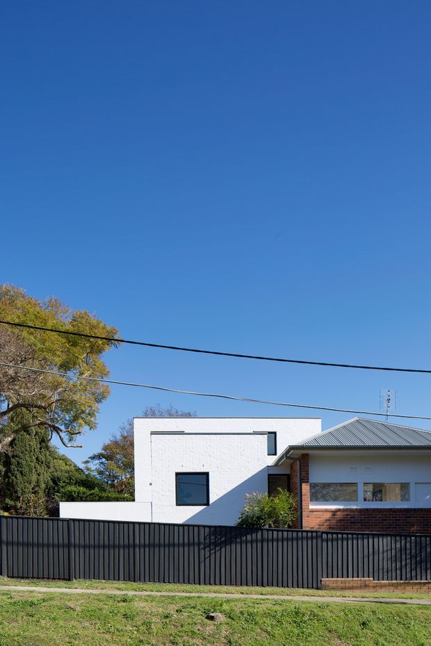 Street-facing view of a low-rise residential house behind a tall dark vertical-panel fence. The dwelling combines a white-painted masonry rectangular volume with a pitched metal-roofed section, exposed red-brown brickwork, and contemporary dark-framed windows. Mature trees, lawn, overhead utility lines, and a clear blue sky surround the property.
