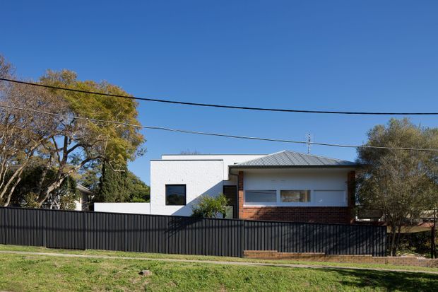 Street-facing view of a low-rise residential house with a contemporary facade. The home combines white rendered or painted masonry, dark brickwork, broad horizontal glazing, and a shallow corrugated metal hipped roof. A tall black metal slat fence encloses the front boundary, with established trees and lawn in the surrounding suburban setting.