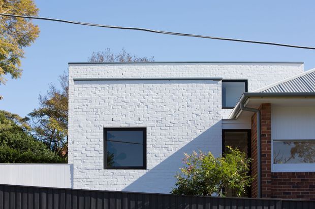 Exterior architectural photograph of a contemporary two-storey residential addition or building volume. The main facade is painted white brick with simple dark-framed rectangular windows, a flat roofline, and a strong angular shadow across the wall. A connected section at right has a pitched corrugated metal roof, red brickwork, and light cladding. A dark vertical fence and established trees frame the foreground and sides.