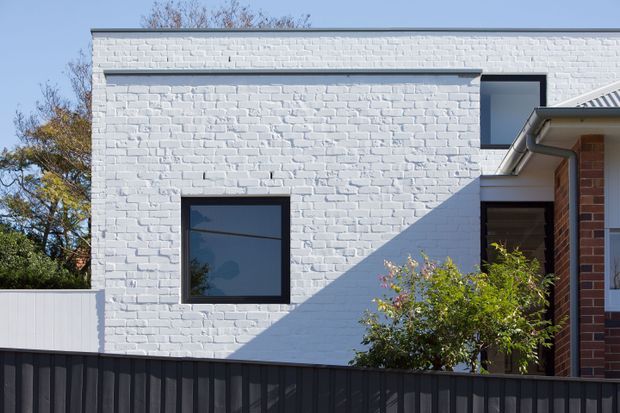 Exterior architectural photograph of a contemporary two-storey residential building. The main facade is clad in painted white brick with simple dark-framed rectangular windows, a projecting upper volume, and a contrasting dark metal boundary fence. A partial pitched roof and red-brick side wall are visible at right, with established trees and shrubs around the property.