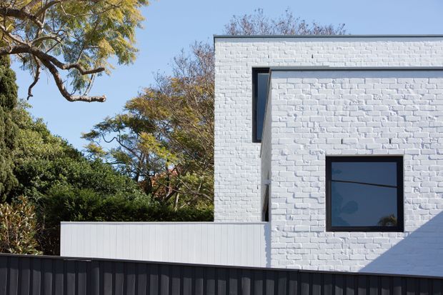 Exterior view of a contemporary white-painted brick building with a simple rectilinear form, dark-framed windows, and a dark corrugated metal boundary fence. Mature trees and dense planting surround the building, while strong sunlight creates sharp geometric shadows across the facade.