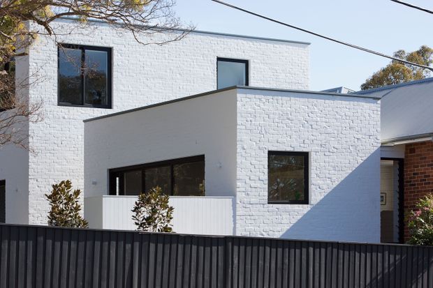 Exterior view of a contemporary multi-level residence with white-painted brick walls, dark-framed windows, a low flat roof, and a projecting box-like front volume. A dark vertical-slat fence and simple landscaping occupy the foreground, with neighbouring residential structures visible at the side.