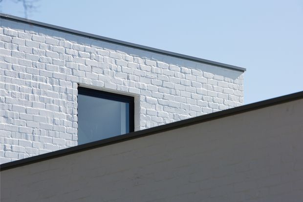A minimalist exterior architectural detail showing two overlapping white-painted brick volumes with dark metal roof and window trim. The foreground wall partially obscures the upper building, which has a flat roofline and a single dark-framed glazed window against a clear blue sky.