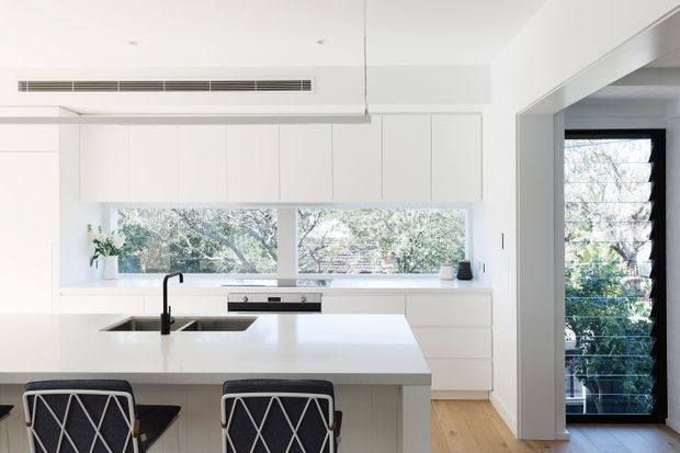 A bright, minimalist contemporary kitchen with extensive white flat-panel cabinetry, a large white island with an integrated black sink and gooseneck tap, and two black-and-white bar stools. A broad horizontal window provides views of trees, while a glazed opening at right leads toward an exterior or light-filled adjoining area. Pale timber flooring and recessed ceiling lighting add warmth to the predominantly white space.