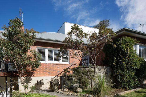 Exterior view of a low-rise brick house with a light grey corrugated metal roof, white-framed louvred windows, and a landscaped front garden with stone edging and retaining walls. The home has a modest stepped form and is partially screened by established shrubs and trees.