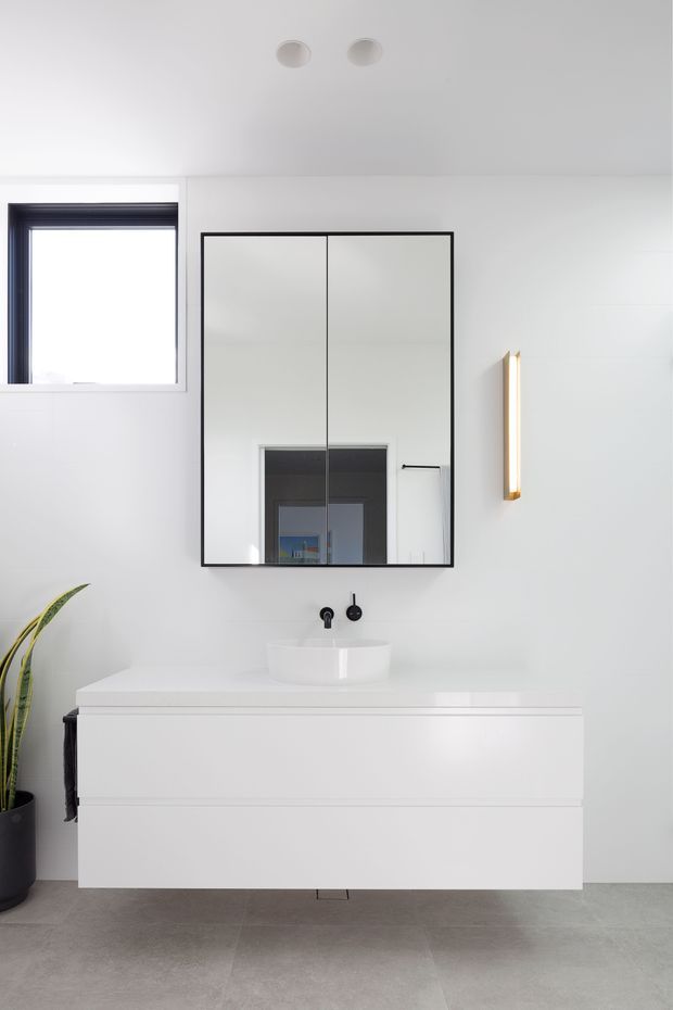 Bright minimalist bathroom vanity with a floating white double-drawer cabinet, white basin, wall-mounted black tapware, and a large black-framed mirrored cabinet. The room has white walls and ceiling, pale grey tiled flooring, a narrow vertical wall light, a high window, and a potted plant.