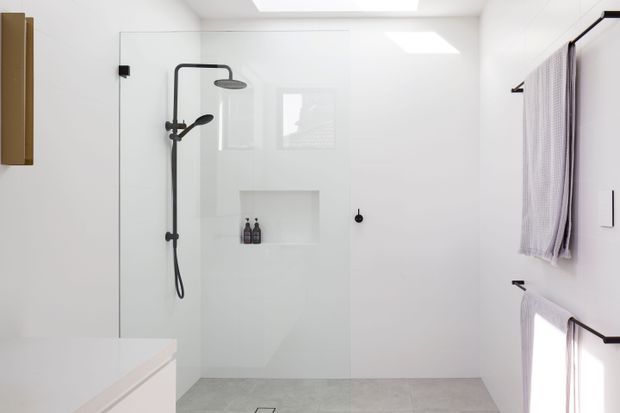 Bright minimalist walk-in bathroom shower with frameless glass screening, matte black shower fittings, a recessed wall niche, pale large-format floor tiles, white walls, a white vanity edge, and wall-mounted towel rails with light towels. A skylight adds natural illumination.