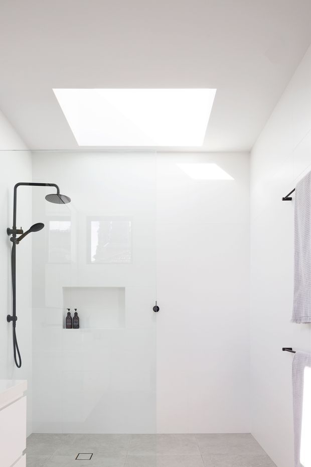 Bright minimalist bathroom with a large frameless glass walk-in shower, matte black shower fittings, recessed shower niche, white tiled walls, large grey floor tiles, skylight, and white towel rails with textured towels.