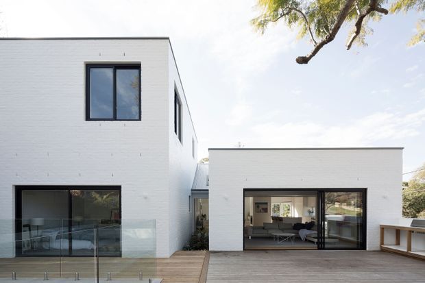 Exterior view of a contemporary white-painted brick residence composed of two clean-lined rectangular volumes. The taller two-storey wing has dark-framed windows and ground-level glazing, while the lower wing opens onto a broad timber deck through a large dark-framed sliding glass door, revealing a furnished living area inside. Glass balustrading, built-in outdoor benching, and mature tree branches are visible around the deck.