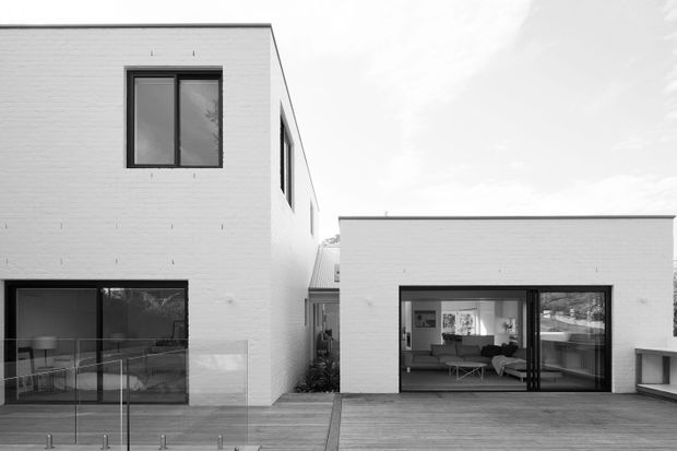 Monochrome exterior view of a modern white-brick, multi-volume residence. The composition shows a taller two-storey wing and a lower flat-roofed living wing connected around a timber deck. Large dark-framed windows and sliding glass doors reveal a contemporary living room inside, while clear glass balustrading lines the foreground deck.