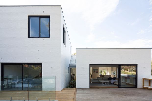 Exterior view of a contemporary white-painted brick residence with simple cubic volumes and flat roofs. The image looks across a timber-clad terrace or deck toward large dark-framed glazed openings, with a balcony and glass balustrade in the foreground. A living room is visible through the open sliding doors.