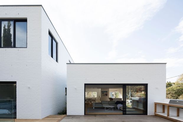 Exterior view of a contemporary white brick residence with crisp cubic volumes, black-framed windows, and a low flat-roofed ground-floor extension opening onto a timber deck. The glazing reveals a living area inside, while the deck includes a built-in outdoor counter or barbecue zone.