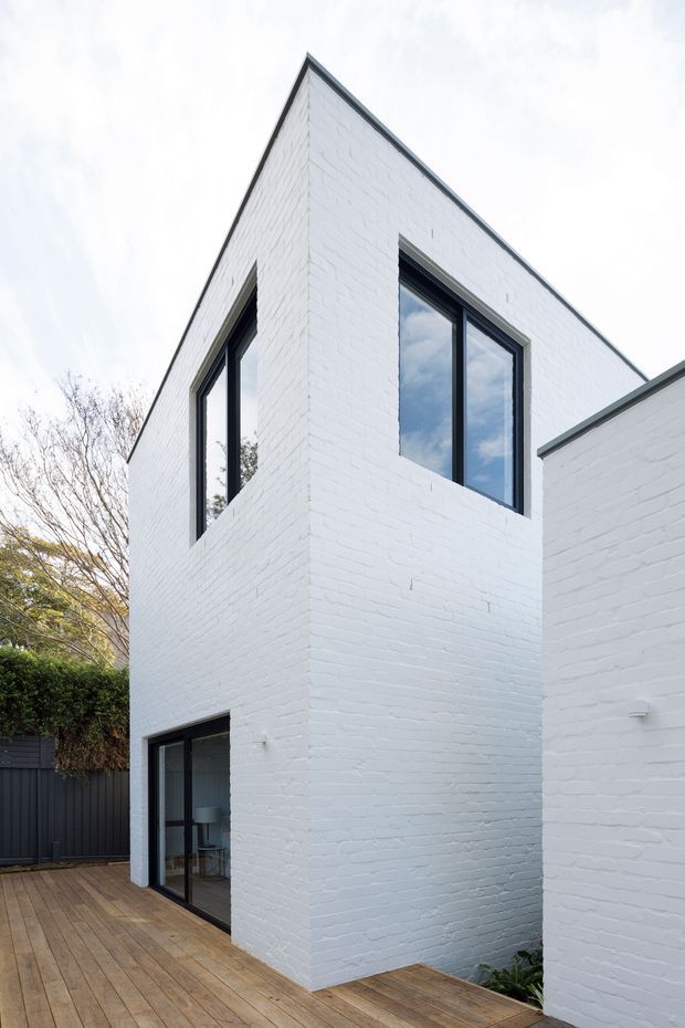 A contemporary two-storey residential extension or house exterior with crisp white-painted brick walls, sharply angled roofline, and dark-framed windows and sliding doors. The building encloses a timber deck courtyard, with mature vegetation and dark fencing visible around the edges. The image emphasizes the clean geometric facade and contrast between white masonry, black window frames, and warm timber decking.